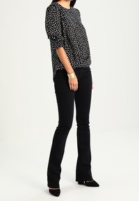 Schwarze Bluse mit weißen Punkten, Rundhalsausschnitt und Dreiviertelärmeln, kombiniert mit schwarzen weit geschnittenen Jeans und spitzen schwarzen Absätzen.