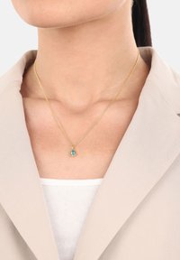 Guldhalsband med en oval blå ädelsten som hänge, på en person klädd i en ljus beige kavaj över en vit topp. Slät kedjedesign.