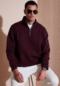 Sudadera burdeos de media cremallera con cuello con cremallera y puños acanalados, combinada con pantalones blancos. Textura suave, diseño minimalista.