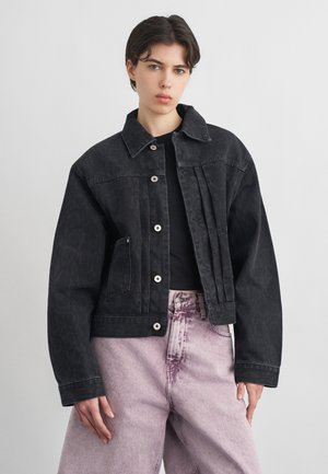 Persona che indossa una giacca di jeans nera con bottoni sopra una maglietta nera e jeans a vita alta color viola chiaro, in piedi davanti a uno sfondo neutro.