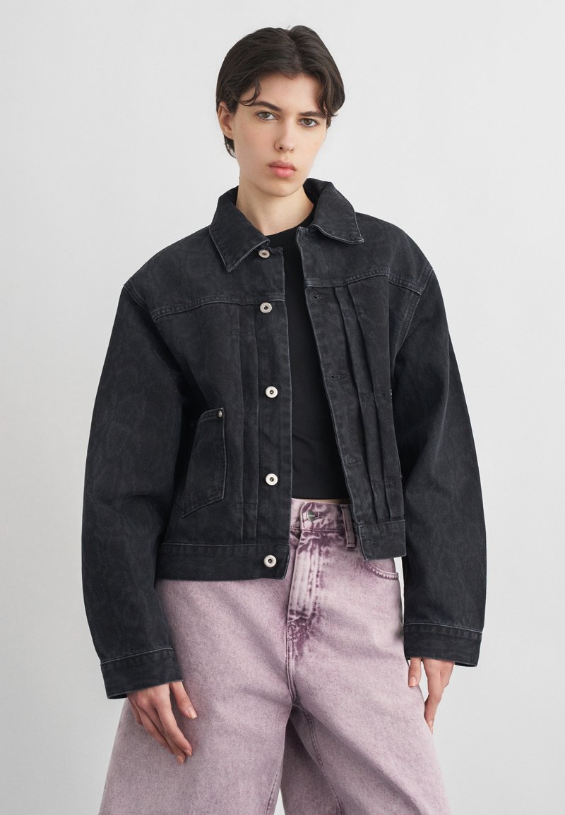 Άτομο που φοράει μαύρο τζιν μπουφάν με κουμπιά πάνω από μαύρο πουκάμισο, με ψηλόμεσα ανοιχτόμωβ τζιν, στέκεται μπροστά σε απλό φόντο.