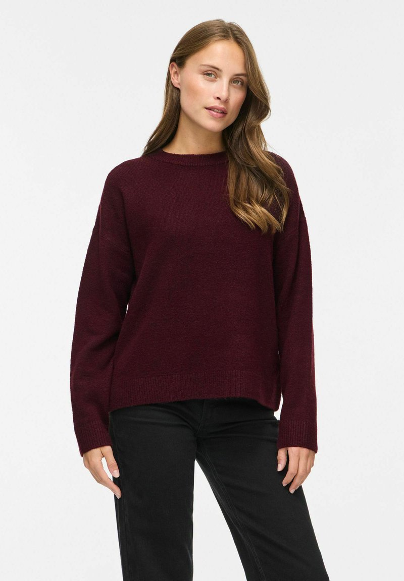 Femme aux longs cheveux châtain clair portant un pull rouge foncé et un pantalon noir sur un fond clair uni.