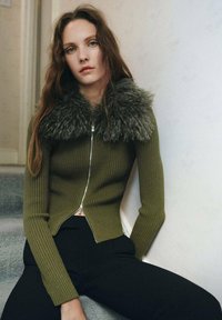 Olive green ribbed knit cardigan with a faux fur collar, featuring a silver zipper and a cropped design paired with black fitted pants.