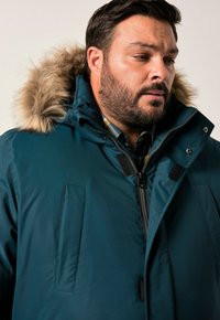 Giacca parka color teal con cappuccio in pelliccia sintetica, zip frontale e due tasche a toppa. Il materiale appare liscio con una finitura opaca.