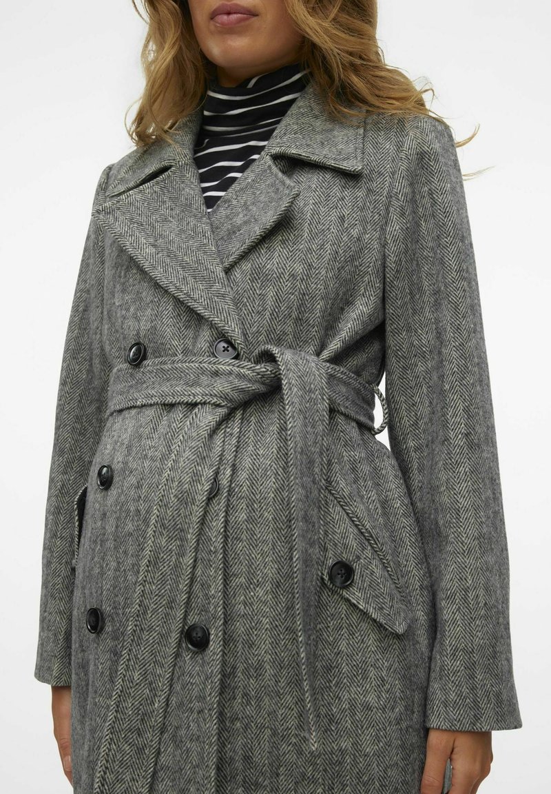 Manteau en chevron gris avec un front à double boutonnage, grandes boutons noirs, et taille ceinturée. Il présente de larges revers et des poches latérales.
