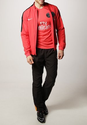 Rote Sportjacke mit schwarzen Akzenten, rotes T-Shirt mit einem Logo, schwarze Hose und schwarze Sportschuhe mit bunten Details.