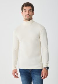 Cămașă cu guler înalt de culoare crem, dintr-un material moale, cu un design ajustat și mâneci lungi. Combinată cu blugi din denim albastru, purtată cu un ceas pe încheietura stângă.