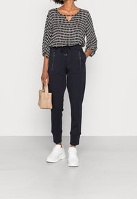 Blouse à motifs noir et blanc avec un col à fente, associée à un pantalon de jogging foncé avec poches zippées et poignets élastiques. Baskets blanches et sac tissé.