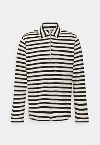 Chemise à manches longues rayée noir et blanc avec col, demi-fermeture éclair à l'avant et une poche poitrine sur fond blanc.