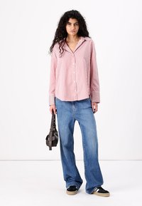 Camisa de manga longa às riscas vermelhas e brancas com botões, combinada com jeans azuis largos e sapatilhas pretas, segurando uma pequena mala escura.