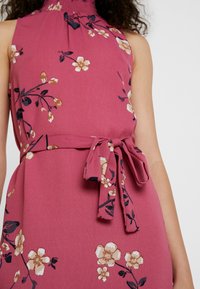 Růžové šaty bez rukávů s béžovými a černými květinovými vzory, stažený výstřih a odpovídající pásek z látky uvázaný do mašle v pase.