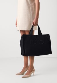 Sac en toile noir avec poignées supérieures, présentant un logo texturé, des œillets en métal et une forme structur ée.