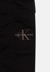 Zwarte stoffen broek met een zijzak en een opvallend geborduurd "CK" logo met daaronder "Calvin Klein Jeans" in metallic draad.