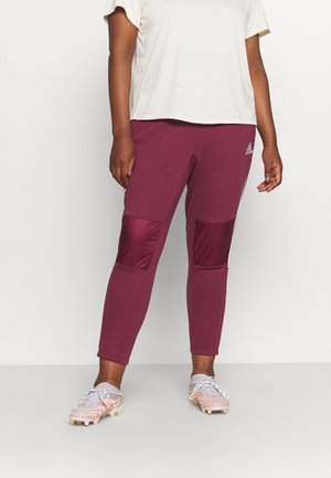 Osoba v maroon sportovních kalhotách Adidas s polstrovanými kolenními záplatami a bílými kopačkami stojí na světlém pozadí.
