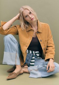Giacca in corduroy senape, canotta marrone, pantaloni wide-leg in denim blu con cintura a strisce bianche e verdi, scarpe slingback marroni, design minimalista.