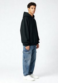 Zwarte capuchonsweatshirt met een zak aan de voorkant, gecombineerd met losse blauwe denim cargobroek met zijzakken en witte sneakers.