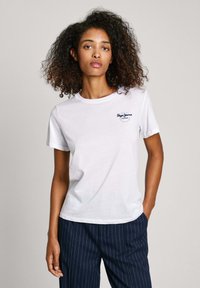 Camiseta blanca de algodón con cuello redondo y mangas cortas; presenta un pequeño logo azul bordado en el lado izquierdo del pecho. Llevada con pantalones a rayas.