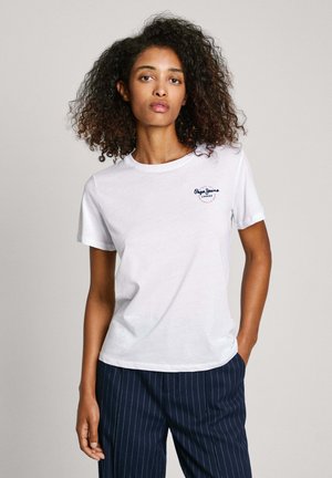 Camiseta blanca de algodón con cuello redondo y mangas cortas; presenta un pequeño logo azul bordado en el lado izquierdo del pecho. Llevada con pantalones a rayas.