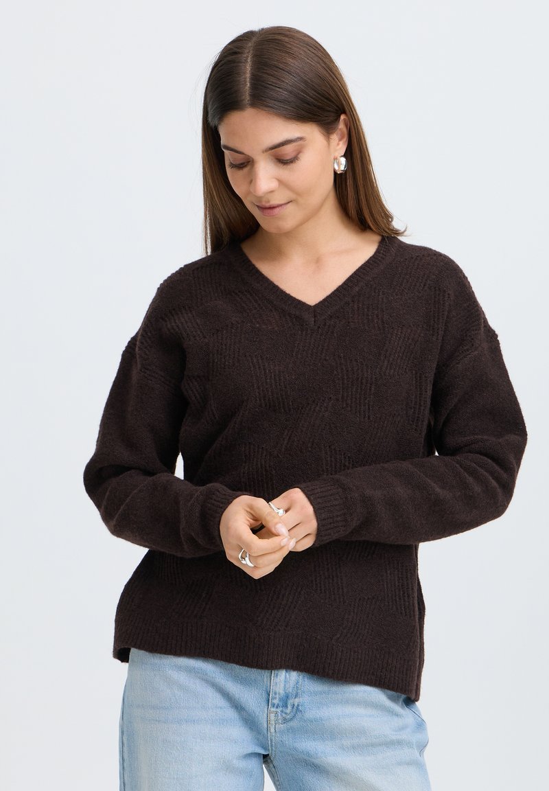Femme aux cheveux bruns et raides portant un pull col en V marron foncé et un jean bleu clair, regardant vers le bas et ajustant des bagues à ses doigts.