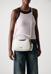 Vit texturerad handväska med en böjd form, guldfärgade detaljer och en avtagbar myntväska. Bärs som crossbody med en justerbar rem.