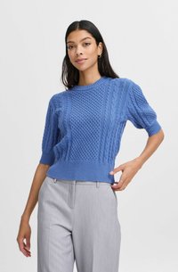 Blauer Strickpullover mit strukturierten Mustern, kurzen Ärmeln und geripptem Saum, kombiniert mit hellgrauen Hosen.