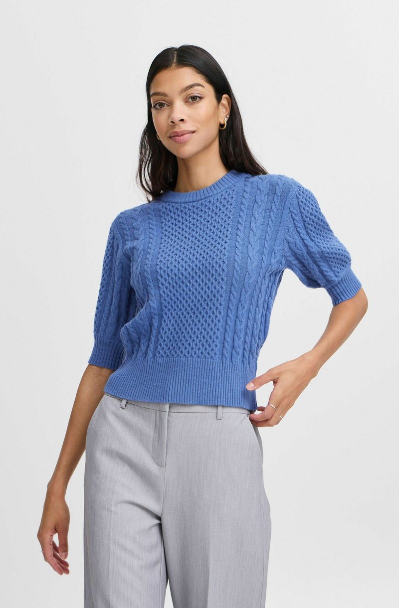 Blauer Strickpullover mit strukturierten Mustern, kurzen Ärmeln und geripptem Saum, kombiniert mit hellgrauen Hosen.