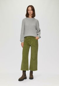 Grauer gestrickter Pullover, olivgrüne Cord-Hose mit weitem Bein, braune Stiefeletten mit strukturierten Sohlen, stehend vor einem neutralen Hintergrund.