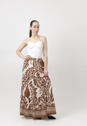 Donna con capelli scuri, indossa una canotta bianca e una lunga gonna marrone e bianca a motivo, in piedi con una mano sui fianchi davanti a uno sfondo semplice.