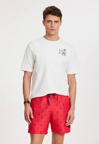 Biały t-shirt z krótkim rękawem z kolorowym nadrukiem na piersi, zestawiony z czerwonymi kąpielówkami z granatowymi motywami roślinnymi i ściągaczem w pasie.