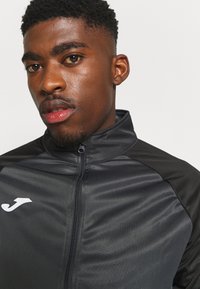 Zwarte sportjack met een ritssluiting aan de voorkant, hoge kraag en contrasterende schouderpanelen. Heeft een wit logo op de borst. Vlotte textuur.