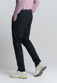 Persoon in zwarte taps toelopende broek en witte sneakers, staand met één hand in de zak tegen een effen achtergrond.