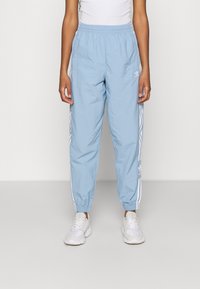 Person som bär ljusblå Adidas träningsoverallsbyxor med vita sidostråk och vita sneakers, stående mot en plain ljus bakgrund.