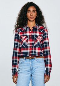 Red, navy, and white plaid flannel shirt with long sleeves, two chest pockets, button-down collar, and a relaxed fit, paired with light blue jeans.
