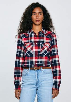 Camicia flanella a quadri rossa, blu navy e bianca, con maniche lunghe, due tasche sul petto, colletto con bottoni e vestibilità morbida, abbinata a jeans blu chiaro.