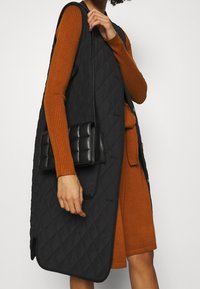 Svart kviltad väst med hög krage, med knäppning fram och sidofickor, bärs över en orange ribbad klänning, kombinerad med en svart handväska.