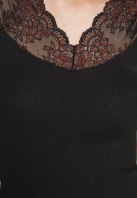 Svart ribbad linne med en genomskinlig spetskrage med blommönster i bränd orange och svart, som framhäver halsområdet.