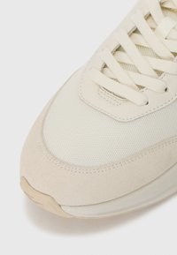 Detailní záběr na béžovou tenisku s špičkou z mesh látky, semišovým překryvem a plochými tkaničkami na bílém pozadí.