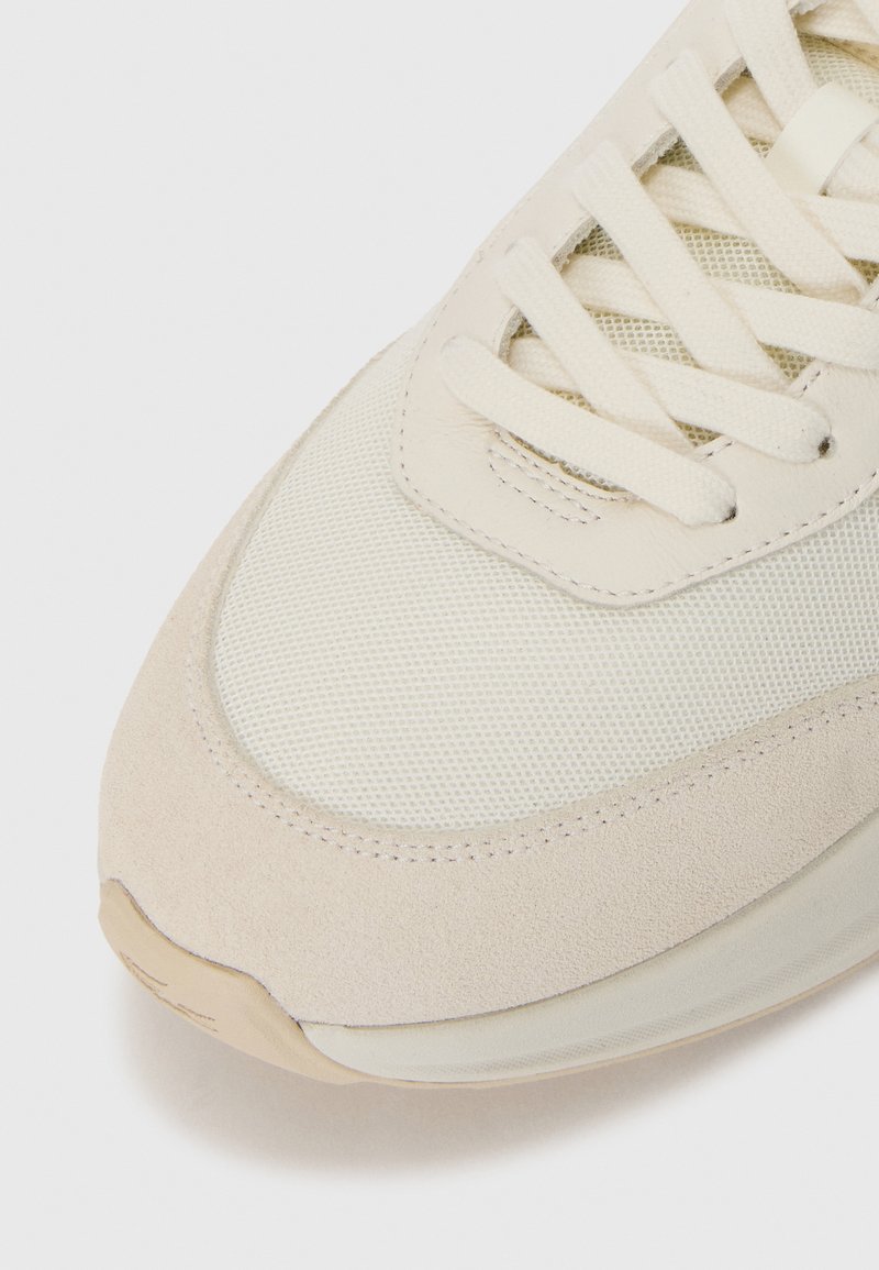Gros plan sur l'orteil d'une sneaker beige avec un tissu en maille, un revêtement en daim et des lacets plats sur un fond blanc.