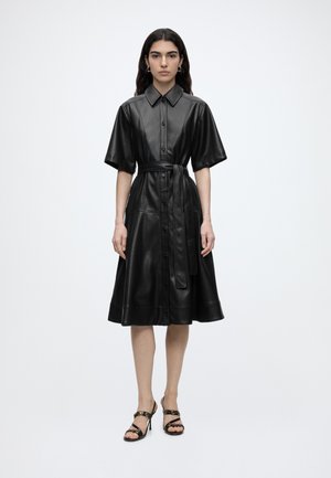 Femme portant une robe en cuir noire à manches courtes boutonnée avec une ceinture à la taille et des sandales noires à talons, debout sur un fond blanc uni.