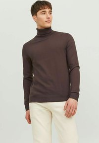 Maglione a collo alto marrone in un design aderente, realizzato in tessuto morbido. Maniche lunghe con polsini a costine, abbinato a pantaloni di colore chiaro.
