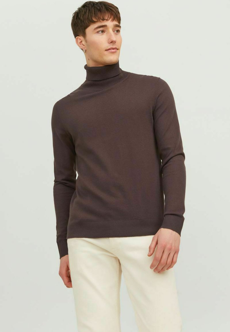 Maglione a collo alto marrone in un design aderente, realizzato in tessuto morbido. Maniche lunghe con polsini a costine, abbinato a pantaloni di colore chiaro.