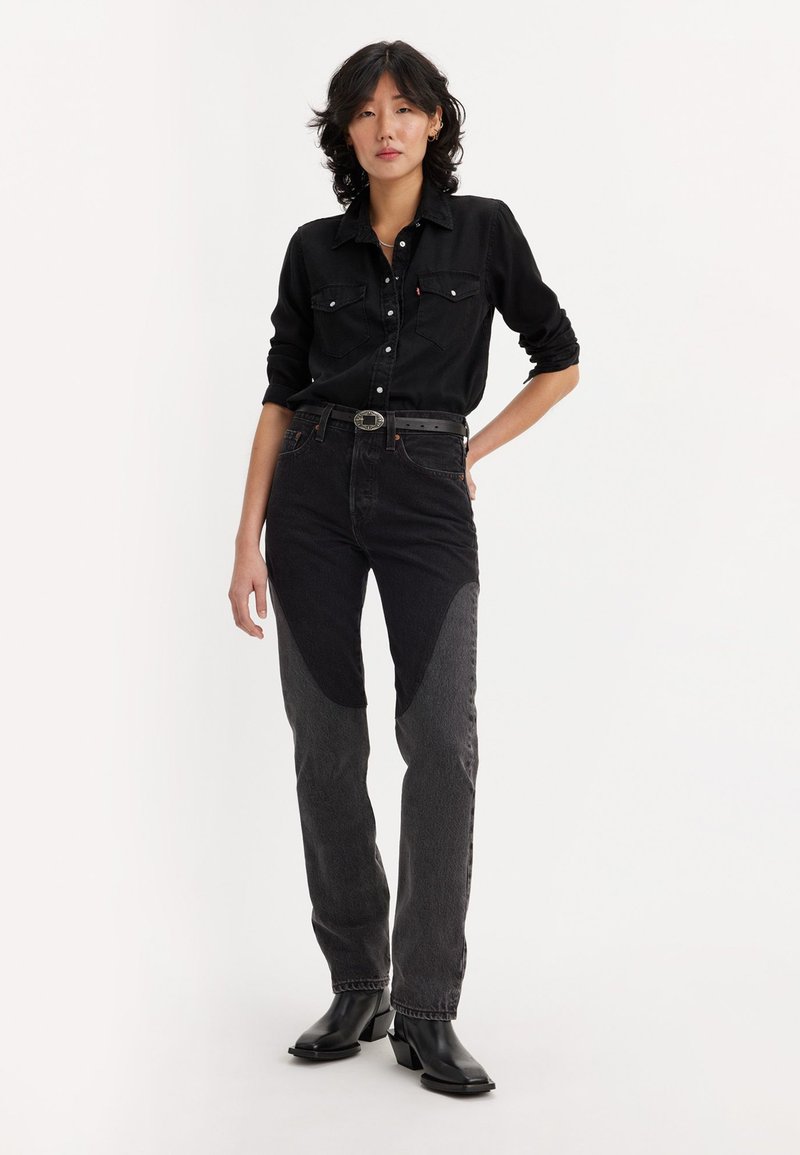 Camisa de mezclilla negra con dos bolsillos en el pecho, combinada con pantalones vaqueros bloqueados en negro y gris que presentan un corte ajustado y una cintura baja. Botas negras hasta el tobillo.