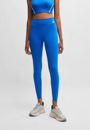 Persoon staande, gekleed in felblauwe legging met hoge taille en bijpassende cropped top, gecombineerd met lichtgrijze sportschoenen.