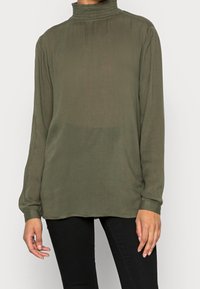 Chemise à manches longues vert olive avec un col haut froncé et une texture subtile. Présente une coupe ample et un ourlet incurvé. Portée sur un pantalon noir.