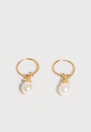 Gouden oorringen met aan elk een enkele witte parel die eraan hangt, weergegeven op een effen lichtgrijze achtergrond.