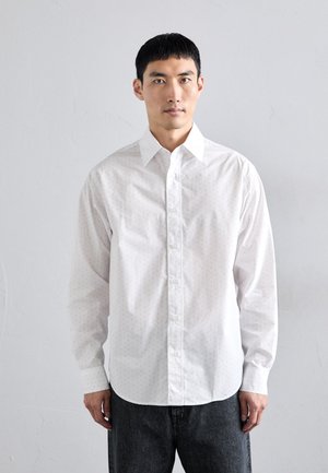 Camisa blanca de botones con un sutil patrón de puntos, de manga larga, con cuello puntiagudo y corte recto, combinada con pantalones de mezclilla oscuros.