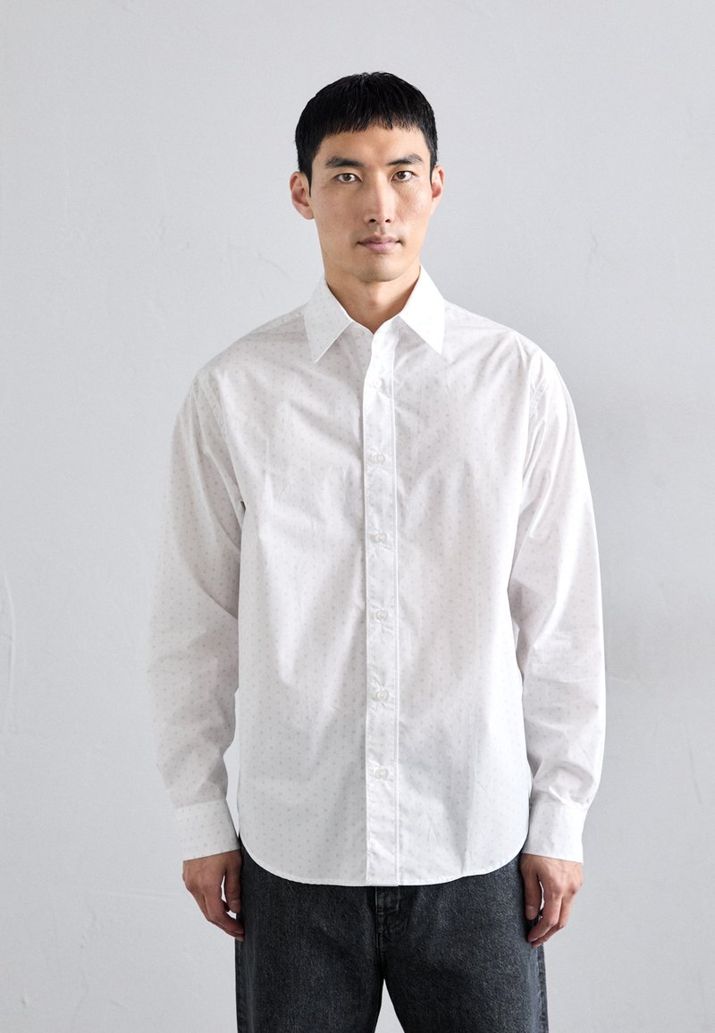 Camisa blanca de botones con un sutil patrón de puntos, de manga larga, con cuello puntiagudo y corte recto, combinada con pantalones de mezclilla oscuros.