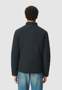 Giacca nera imbottita con colletto alto, caratterizzata da una vestibilità dritta e cuciture visibili. Indossata sopra jeans azzurri chiari, con un minimo dettaglio di branding.