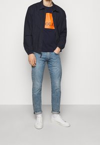 Marinblå bomberjacka över en marinblå och orange grafisk T-shirt, ljusblå jeans och vita sneakers med upprullade cuffs.
