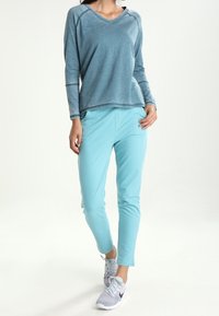 Jasnoniebieska bluzka z długim rękawem, dekoltem w serek i rękawami raglanowymi, zestawiona z dopasowanymi, zwężanymi spodniami. Modelka ma na sobie szare buty sportowe.